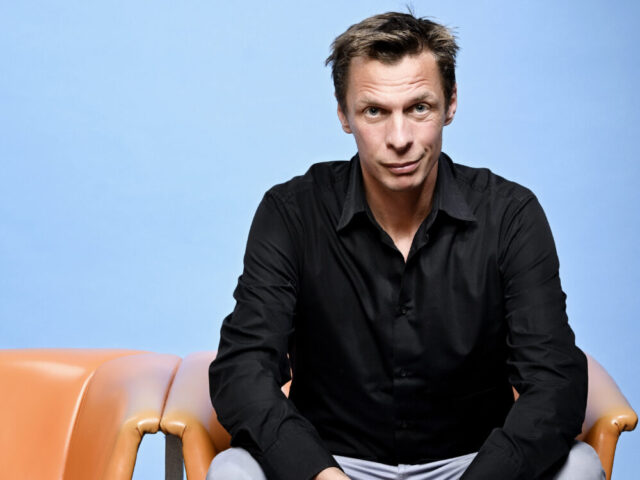 Klaus Eckel sitzt auf einem orangen Stuhl vor blauem Hintergrund