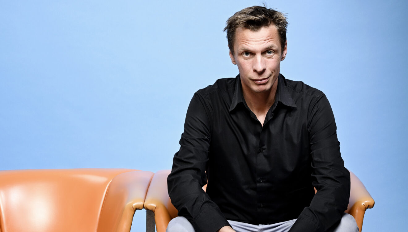 Klaus Eckel sitzt auf einem orangen Stuhl vor blauem Hintergrund